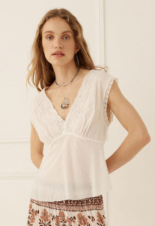 a-woman-wearing-a-white-sleeveless-lace-trimmed-v-neck-top-with-a-gathered-waist-paired-with-a-brown-and-cream-patterned-skirt-standing-against-a-white-paneled-wall