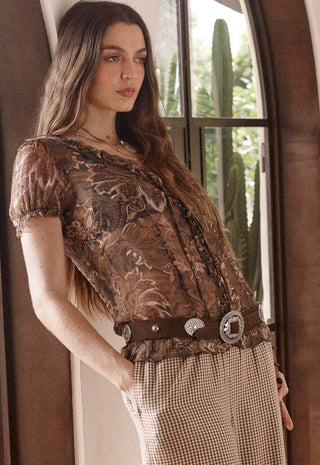 a-woman-wearing-a-brown-floral-lace-top-and-a-brown-patterned-skirt-accessorized-with-a-wide-brown-belt-featuring-silver-conchos-standing-in-front-of-a-window-with-cacti-visible-outside
