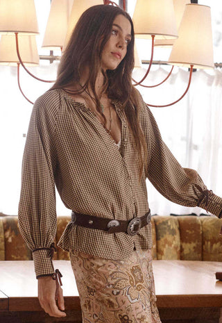 a-woman-wearing-a-brown-and-beige-gingham-long-sleeve-blouse-with-a-dark-brown-belt-and-a-matching-patterned-skirt-standing-in-a-warmly-lit-indoor-setting-with-a-chandelier-and-upholstered-bench-in-the-background