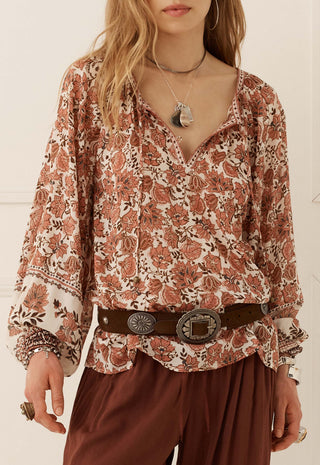a-woman-wearing-a-long-sleeve-bohemian-style-blouse-with-a-rust-and-brown-floral-paisley-print-on-a-cream-background-styled-with-a-dark-brown-belt-and-brown-pants