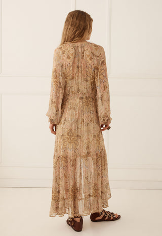 a-woman-viewed-from-behind-wearing-a-long-sleeved-midi-length-beige-maxi-dress-with-an-intricate-paisley-pattern-in-warm-brown-and-tan-tones-she-is-wearing-brown-strappy-flat-sandals-against-a-clean-white-paneled-wall-background