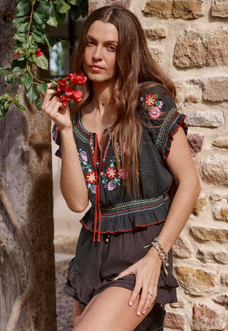 a-woman-wearing-a-dark-gray-embroidered-blouse-and-matching-shorts-stands-against-a-rustic-stone-wall-holding-red-flowers