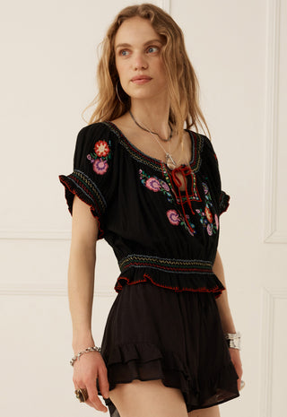 a-woman-wearing-a-black-embroidered-short-sleeve-top-and-matching-black-ruffled-shorts-standing-against-a-white-paneled-wall
