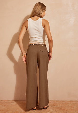 a-woman-shown-from-the-back-wearing-wide-leg-brown-trousers-and-a-white-ribbed-tank-top-standing-against-a-textured-beige-wall