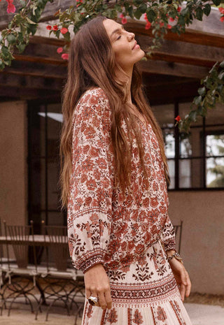a-woman-wearing-a-bohemian-style-long-sleeve-dress-with-a-rust-brown-and-cream-floral-pattern-standing-in-an-outdoor-patio-setting-with-a-rustic-wooden-structure-and-hanging-greenery-in-the-background