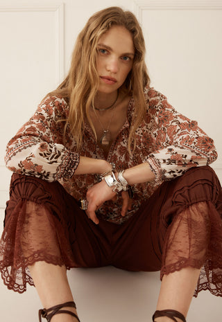 a-woman-wearing-a-bohemian-style-brown-and-beige-patterned-blouse-with-sheer-lace-trimmed-sleeves-and-dark-brown-ruffled-pants-seated-against-a-white-paneled-wall