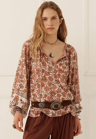 a-woman-modeling-a-long-sleeve-bohemian-style-blouse-with-a-rust-brown-and-cream-floral-pattern-paired-with-a-dark-brown-belt-and-brown-pants-set-against-a-white-paneled-wall