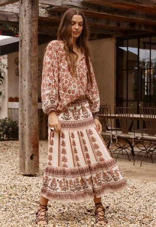 a-woman-wearing-a-bohemian-style-two-piece-dress-set-with-a-rust-brown-and-cream-floral-print-standing-on-a-gravel-patio-in-front-of-a-rustic-wooden-structure