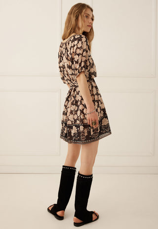 a-woman-wearing-a-short-black-and-beige-floral-print-dress-styled-with-black-knee-high-studded-sandals-standing-against-a-white-paneled-wall