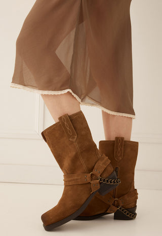 a-pair-of-brown-suede-ankle-boots-with-decorative-straps-and-chain-details-shown-on-a-model-wearing-sheer-brown-draped-pants-against-a-light-cream-background