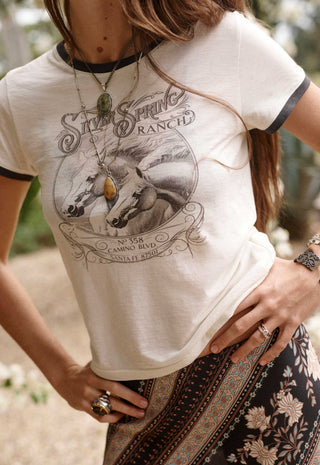 a-woman-wearing-a-cream-colored-ringer-t-shirt-with-black-trim-and-a-vintage-silk-spring-ranch-graphic-print-on-the-front-paired-with-a-dark-patterned-skirt