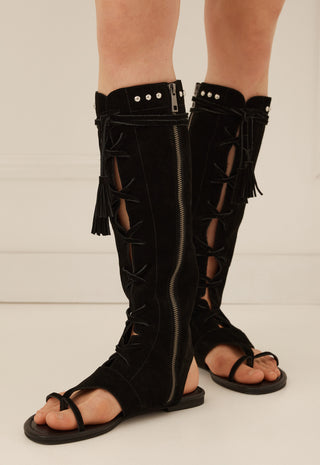 black-suede-knee-high-strappy-sandals-with-side-zippers-cutout-details-and-fringe-shown-on-a-models-feet-against-a-light-neutral-background