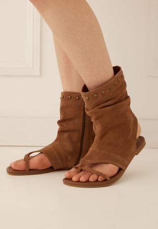 a-pair-of-brown-suede-slide-sandals-with-wraparound-ankle-straps-and-metal-studs-shown-on-a-models-feet-against-a-light-cream-background